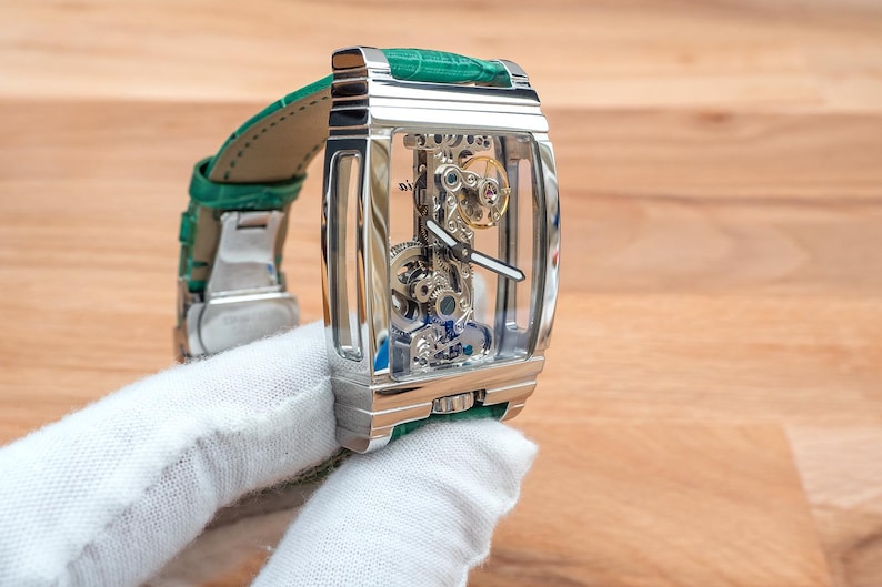 May include: A silver rectangular wristwatch with a transparent face, displaying the internal mechanisms. It features a green leather strap. The watch is held by a gloved hand.
