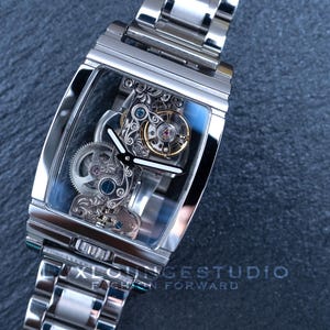 Puede incluir: Un reloj esqueleto plateado con una esfera transparente y una correa de metal plateado. La esfera del reloj muestra el intrincado funcionamiento interno del reloj.