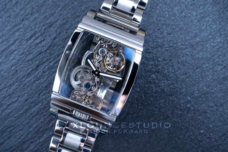 Puede incluir: Un reloj esqueleto plateado con una esfera transparente y una correa de metal plateado. La esfera del reloj muestra el intrincado funcionamiento interno del reloj.