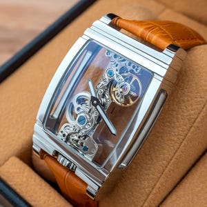 May include: A rectangular, silver-toned wristwatch with a transparent face, showcasing intricate internal mechanisms. The watch has a brown leather band and is presented in a brown box. The watch face displays silver hands and blue and gold accents.