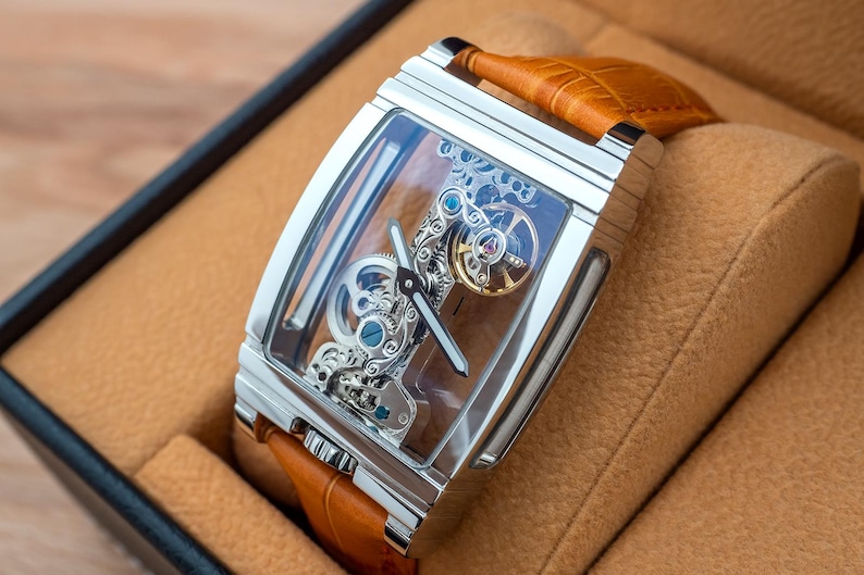 May include: A rectangular, silver-toned wristwatch with a transparent face, showcasing intricate internal mechanisms. The watch has a brown leather band and is presented in a brown box. The watch face displays silver hands and blue and gold accents.
