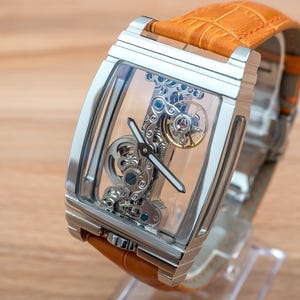 May include: A rectangular, silver-toned wristwatch with a transparent face, showcasing the intricate internal mechanisms. The watch has a tan leather band. The watch is displayed on a clear stand against a light brown wooden surface.