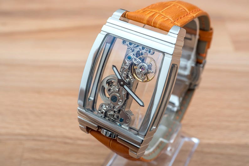 May include: A rectangular, silver-toned wristwatch with a transparent face, showcasing the intricate internal mechanisms. The watch has a tan leather band. The watch is displayed on a clear stand against a light brown wooden surface.