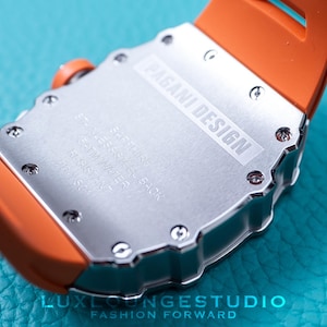 Peut inclure: Gros plan d'une montre Pagani Design. La montre a un dos argent&eacute; avec le nom de la marque et les sp&eacute;cifications. Elle est dot&eacute;e d'un bracelet en silicone orange. La montre est sur une surface turquoise.