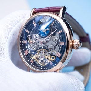 May include: A rose gold skeleton watch with a black face and Roman numeral hour markers. The watch features a moon phase complication and a visible mechanical movement. The watch is on a brown leather strap.