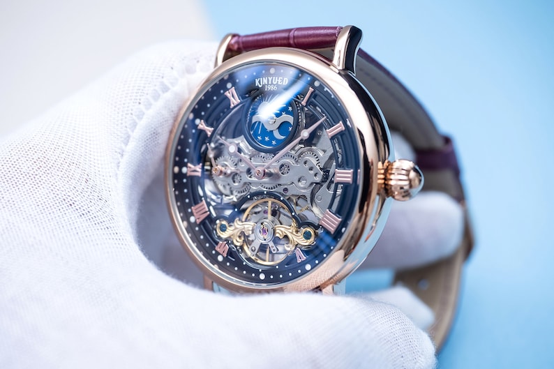 May include: A rose gold skeleton watch with a black face and Roman numeral hour markers. The watch features a moon phase complication and a visible mechanical movement. The watch is on a brown leather strap.