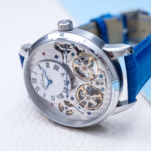 May include: A silver-toned wristwatch with a white face and blue hands. The watch has a skeletonized design, revealing the intricate inner workings of the movement. The watch is on a blue leather strap.