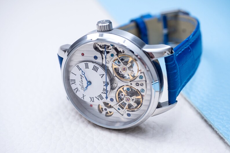 May include: A silver-toned wristwatch with a white face and blue hands. The watch has a skeletonized design, revealing the intricate inner workings of the movement. The watch is on a blue leather strap.