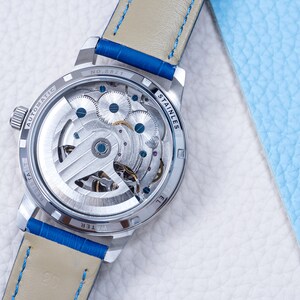 May include: The back of a silver watch with a see-through case, revealing the intricate inner workings of the automatic movement. The watch has a blue leather strap with white stitching and the words "WATER RESIST" and "STAINLESS STEEL" are visible on the case.