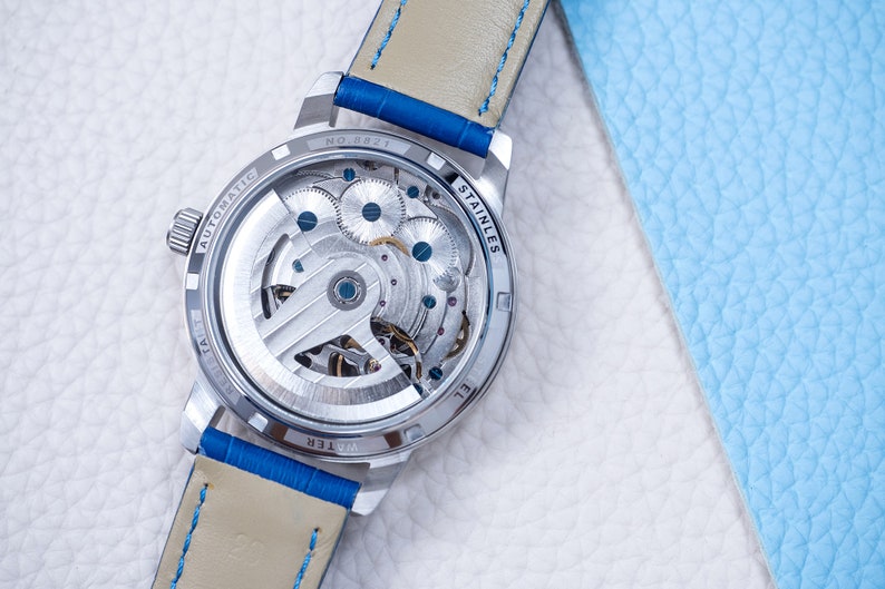 May include: The back of a silver watch with a see-through case, revealing the intricate inner workings of the automatic movement. The watch has a blue leather strap with white stitching and the words "WATER RESIST" and "STAINLESS STEEL" are visible on the case.