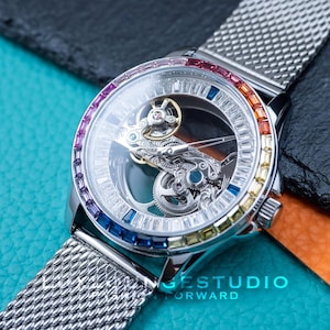 Könnte beinhalten: Eine silberne Uhr mit einem skelettierten Zifferblatt und einem silbernen Mesh-Armband. Das Zifferblatt der Uhr ist von einer Reihe bunter Edelsteine umgeben.