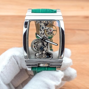 May include: A rectangular, silver wristwatch with a transparent face, revealing the internal mechanisms. It has a green leather strap. The watch is held by a gloved hand, against a wooden background.