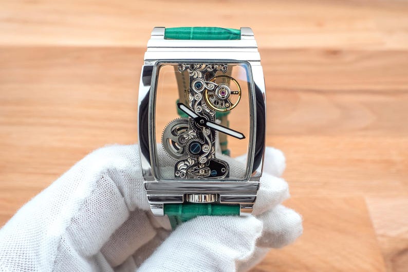 May include: A rectangular, silver wristwatch with a transparent face, revealing the internal mechanisms. It has a green leather strap. The watch is held by a gloved hand, against a wooden background.