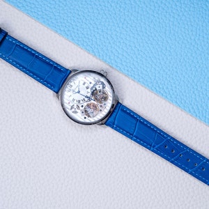 May include: A silver skeleton watch with a blue leather band. The watch face has Roman numerals and a visible gear mechanism.