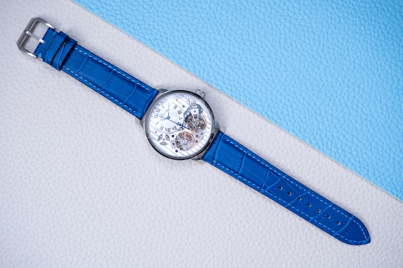 May include: A silver skeleton watch with a blue leather band. The watch face has Roman numerals and a visible gear mechanism.