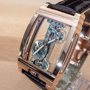 May include: A rose gold rectangular wristwatch with a transparent face, showcasing the intricate internal mechanisms. The watch has a black leather band and is displayed on a clear stand. The watch's internal components are silver and blue.