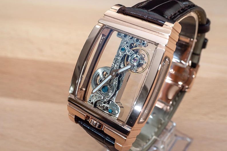 May include: A rose gold rectangular wristwatch with a transparent face, showcasing the intricate internal mechanisms. The watch has a black leather band and is displayed on a clear stand. The watch's internal components are silver and blue.
