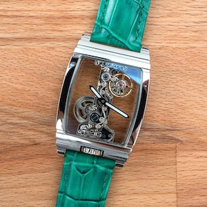 May include: A rectangular, silver wristwatch with a transparent face, revealing intricate internal mechanisms. The watch has a green leather strap. The watch is displayed on a wooden surface.