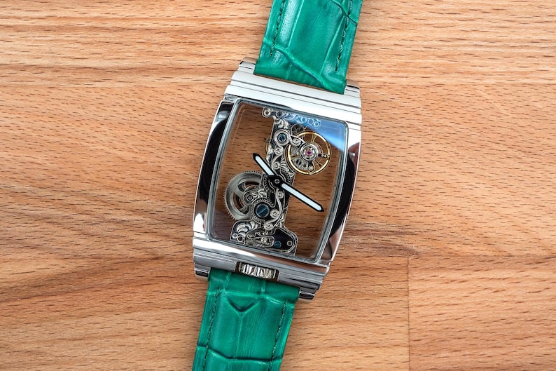 May include: A rectangular, silver wristwatch with a transparent face, revealing intricate internal mechanisms. The watch has a green leather strap. The watch is displayed on a wooden surface.