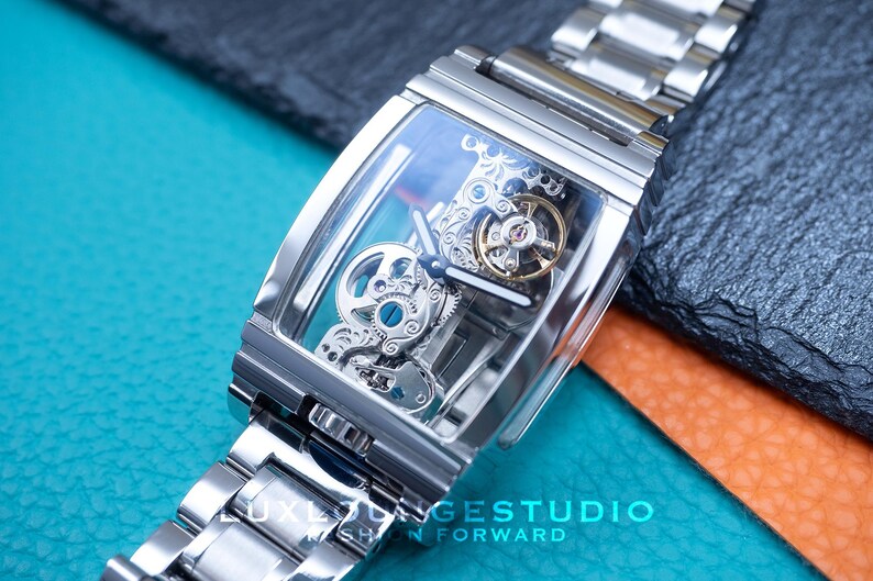 Puede incluir: Un reloj plateado con una esfera transparente y un movimiento mec&aacute;nico visible. El reloj tiene una caja rectangular y una correa de metal plateado.