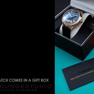 Puede incluir: Una caja de regalo negra con un reloj dorado en su interior. El reloj tiene una esfera azul con agujas doradas y n&uacute;meros romanos. El reloj est&aacute; en una correa de cuero negra. La caja est&aacute; sobre una superficie turquesa con una tarjeta de visita negra que dice "LuxLoungeStudio" y una direcci&oacute;n de sitio web.