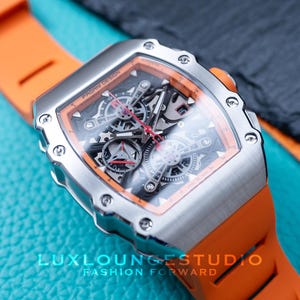 Peut inclure: Une montre-bracelet argent&eacute;e avec un bracelet orange. Le cadran de la montre est transparent, r&eacute;v&eacute;lant les m&eacute;canismes internes. La montre est pos&eacute;e sur un fond bleu et noir. Le texte "PAGANI DESIGN" est visible sur le cadran.