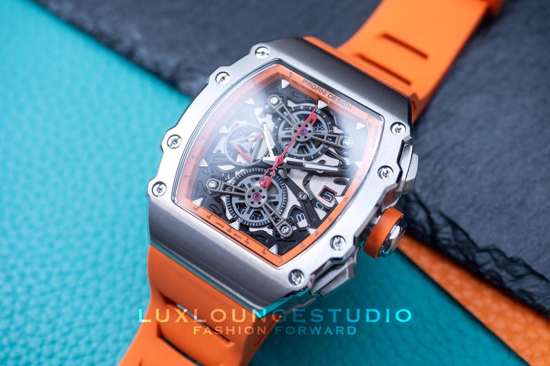 Peut inclure: Une montre argent&eacute;e avec un bracelet orange. Le cadran de la montre pr&eacute;sente un design complexe avec des engrenages visibles et une palette de couleurs noire et orange. C'est un article de luxe.