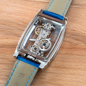 May include: A silver watch with a clear face and a blue leather band. The watch face shows the intricate inner workings of the watch. The band has a blue stitching detail. The watch is on a wooden surface.