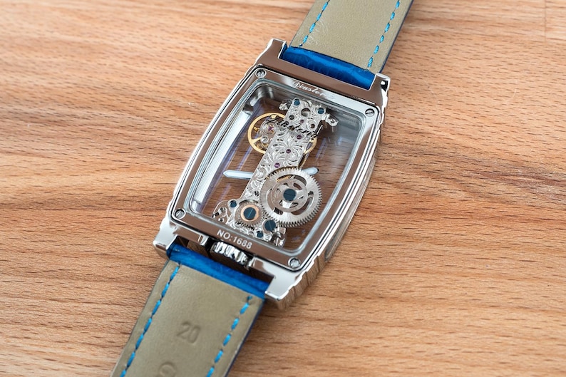 May include: A silver watch with a clear face and a blue leather band. The watch face shows the intricate inner workings of the watch. The band has a blue stitching detail. The watch is on a wooden surface.