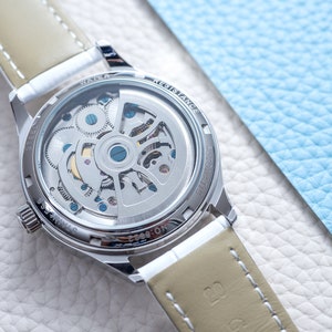 Pu&ograve; includere: Un orologio argentato con un cinturino in pelle marrone. L'orologio ha un fondello trasparente che mostra il meccanismo interno dell'orologio. Il testo "WATER RESISTANCE" &egrave; visibile sul retro dell'orologio.