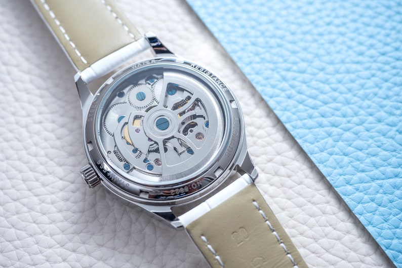 Pu&ograve; includere: Un orologio argentato con un cinturino in pelle marrone. L'orologio ha un fondello trasparente che mostra il meccanismo interno dell'orologio. Il testo "WATER RESISTANCE" &egrave; visibile sul retro dell'orologio.