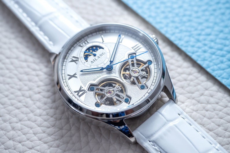 Pu&ograve; includere: Un orologio d'argento con quadrante bianco e lancette blu. L'orologio ha una complicazione di fase lunare e un design a cuore aperto, che rivela i meccanismi intricati del movimento. L'orologio &egrave; su un cinturino in pelle bianca.