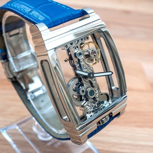 May include: A silver skeleton watch with a blue leather band. The watch face is transparent, revealing the intricate inner workings of the watch.