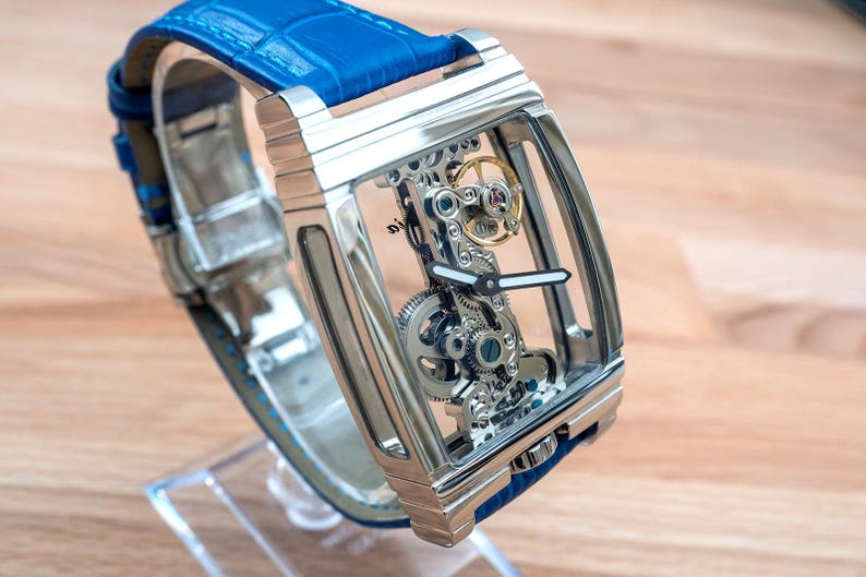 May include: A silver skeleton watch with a blue leather band. The watch face is transparent, revealing the intricate inner workings of the watch.