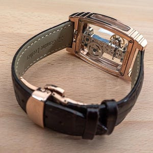 May include: A rose gold-toned wristwatch with a transparent face, showcasing the intricate internal mechanisms. The watch features a black leather strap with a rose gold clasp. The words "Genuine Leather" are visible on the strap.