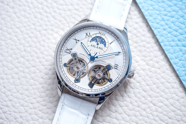 Pu&ograve; includere: Un orologio da polso bianco con quadrante argentato e cinturino in pelle bianca. L'orologio ha un tourbillon a doppia volata e una complicazione di fase lunare. Il quadrante dell'orologio presenta anche il testo "AILANG" e "DOUBLE FLY TOURBILLON 21 JEWELS AUTOMATIC".