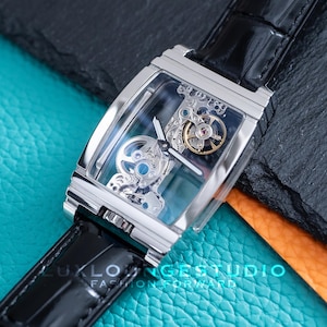 Könnte beinhalten: Eine silberne Uhr mit einem schwarzen Lederarmband. Das Zifferblatt der Uhr ist transparent und zeigt das Innenleben der Uhr.