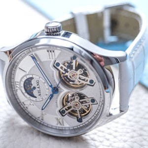 Pu&ograve; includere: Un orologio da polso argentato con quadrante bianco e indici delle ore in numeri romani. L'orologio ha un indicatore di fase lunare, una finestrella per la data e un design a cuore aperto che rivela i complessi meccanismi dell'orologio. L'orologio &egrave; su un cinturino in pelle bianca.