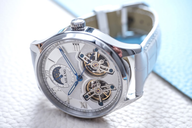 Pu&ograve; includere: Un orologio da polso argentato con quadrante bianco e indici delle ore in numeri romani. L'orologio ha un indicatore di fase lunare, una finestrella per la data e un design a cuore aperto che rivela i complessi meccanismi dell'orologio. L'orologio &egrave; su un cinturino in pelle bianca.