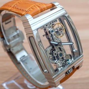 May include: A rectangular, skeletonized wristwatch with a silver-tone case and a tan leather strap. The watch face reveals the intricate inner workings of the mechanism. The strap is made of textured leather. The watch is displayed on a clear stand.