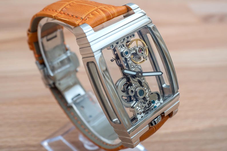 May include: A rectangular, skeletonized wristwatch with a silver-tone case and a tan leather strap. The watch face reveals the intricate inner workings of the mechanism. The strap is made of textured leather. The watch is displayed on a clear stand.
