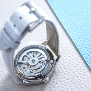 Pu&ograve; includere: Un orologio argentato con un cinturino in pelle bianca. Il retro dell'orologio &egrave; trasparente, mostrando il complesso meccanismo interno dell'orologio.