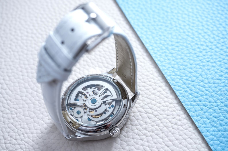 Pu&ograve; includere: Un orologio argentato con un cinturino in pelle bianca. Il retro dell'orologio &egrave; trasparente, mostrando il complesso meccanismo interno dell'orologio.