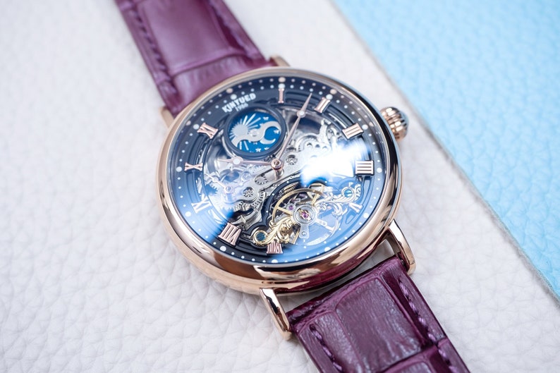 May include: A rose gold watch with a burgundy leather strap. The watch face is a skeleton design with a moon phase complication and a black and blue dial. The watch face has Roman numeral hour markers.