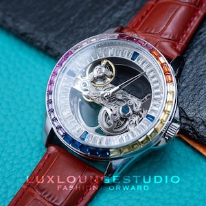 May include: A silver skeleton watch with a clear face and a brown leather strap. The watch face is surrounded by a row of colourful gemstones.