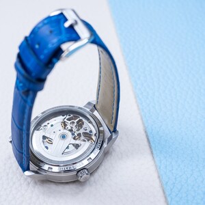 May include: A silver watch with a blue leather band. The watch has a clear back that shows the inner workings of the watch.