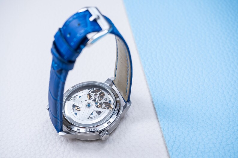 May include: A silver watch with a blue leather band. The watch has a clear back that shows the inner workings of the watch.