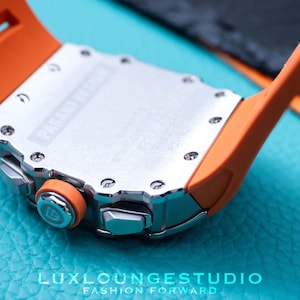 Peut inclure: Gros plan d'une montre de luxe avec un bracelet orange et un bo&icirc;tier argent&eacute;. La montre pr&eacute;sente un design d&eacute;taill&eacute; avec des vis visibles et le texte "PAGANI DESIGN". La montre est pos&eacute;e sur un fond turquoise.