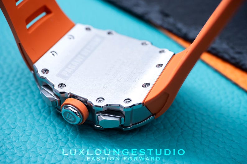 Peut inclure: Gros plan d'une montre de luxe avec un bracelet orange et un bo&icirc;tier argent&eacute;. La montre pr&eacute;sente un design d&eacute;taill&eacute; avec des vis visibles et le texte "PAGANI DESIGN". La montre est pos&eacute;e sur un fond turquoise.