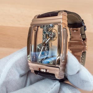 May include: A rose gold rectangular wristwatch with a transparent face, showcasing the intricate internal mechanisms. The watch has a brown leather strap and is held by a gloved hand. The watch's design is modern and luxurious.